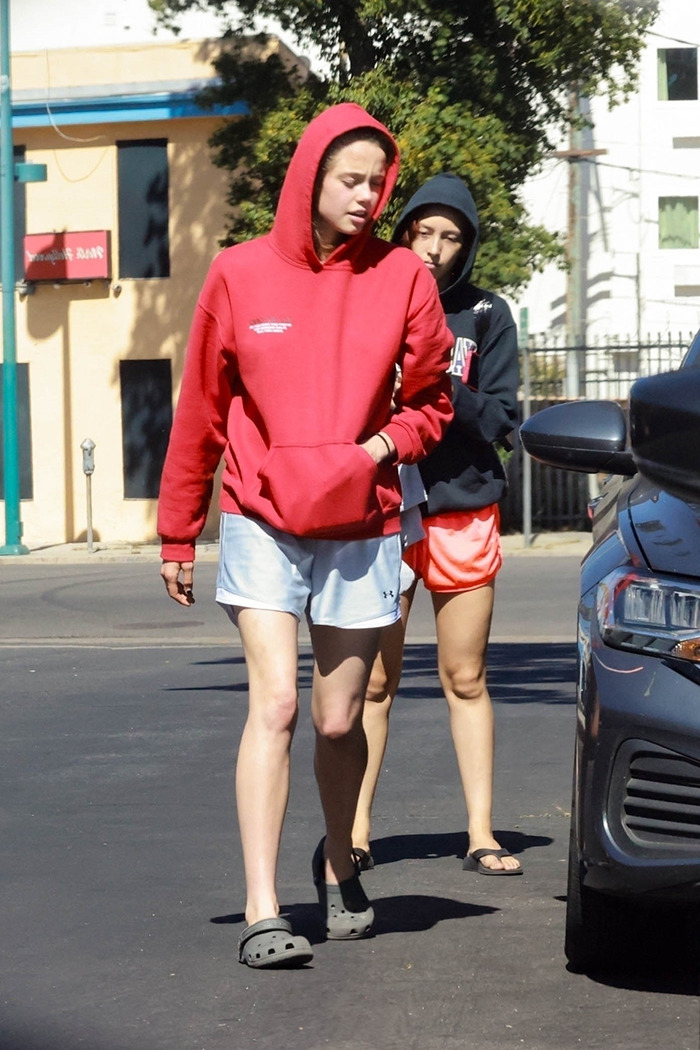 Shiloh Jolie-Pitt, 19, wearing a red hoodie and shorts, walking outdoors with a companion in casual attire.
