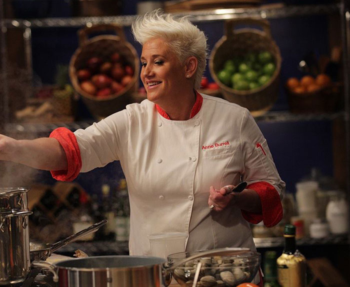 Chef Anne Burrell cooking in a professional kitchen surrounded by fresh produce and kitchenware, honoring stars we've lost.