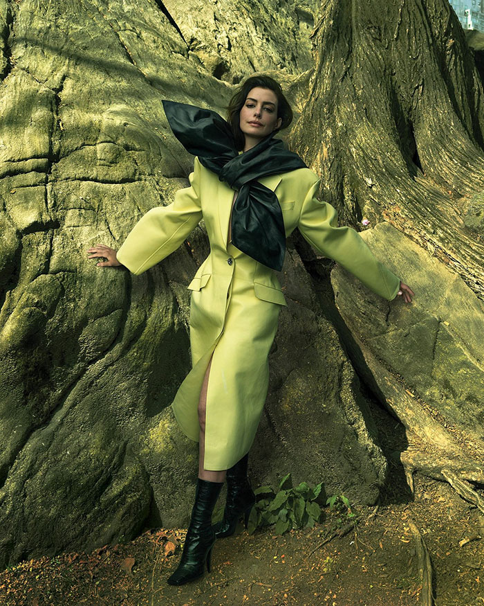 Anne Hathaway posing against a large tree in a yellow coat and black boots during a Vogue photoshoot. Anne Hathaway posing against a large tree in a yellow coat and black boots during a Vogue photoshoot.