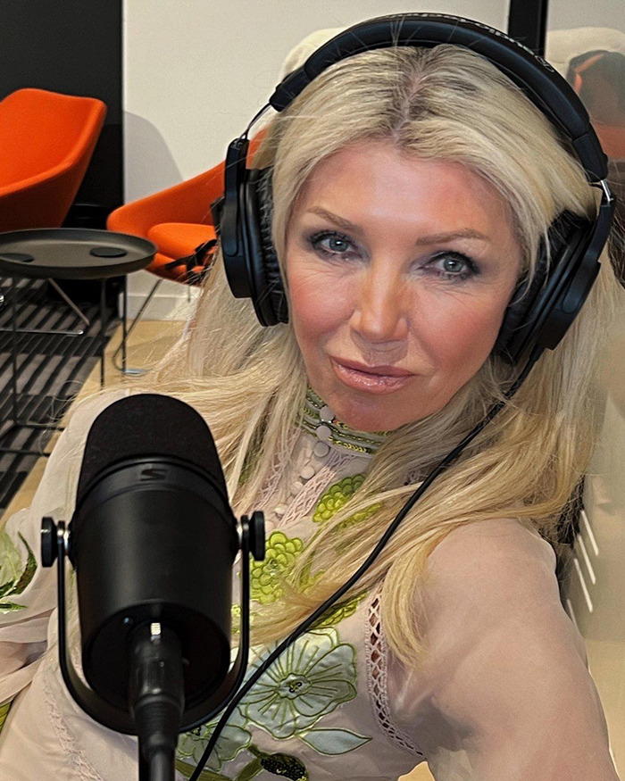 Blonde woman wearing headphones speaking into a microphone in a podcast studio with orange chairs in the background.
