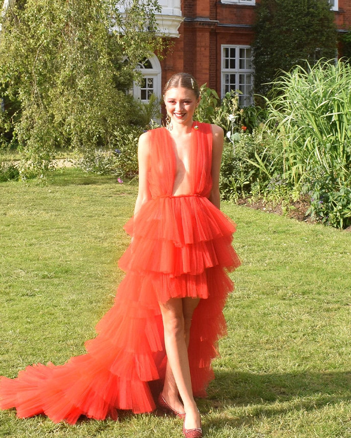 Young woman in a vibrant red tiered dress posing outdoors in a garden setting, representing 23YO anti-vax woman story.