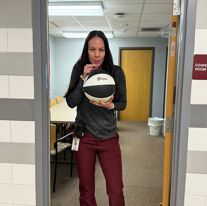 Woman creatively sipping through a straw from a basketball inside an office hallway, showcasing unique drinking ideas.