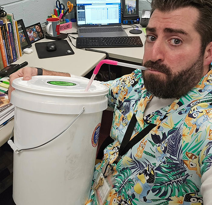 Man wearing a colorful shirt drinking with a straw from a large white bucket at his office desk creative sippers idea