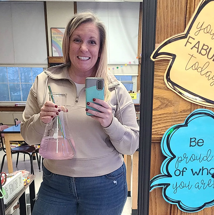 Woman holding a science beaker with a straw, drinking from an unconventional sipper in a classroom setting.