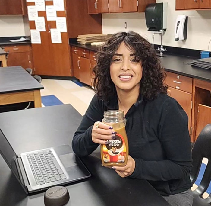 Woman sitting at desk holding a creative sipper using a Nescafe container instead of a cup in a classroom setting