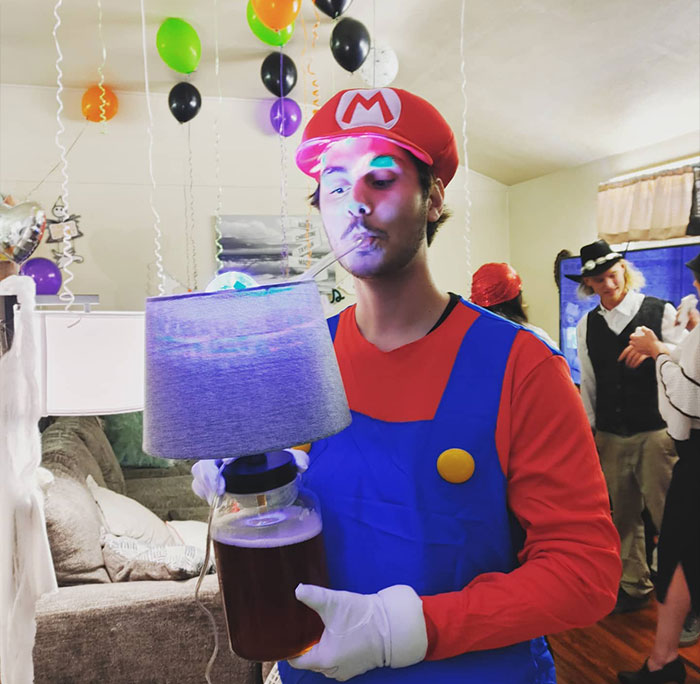Person dressed as Mario sipping a drink through a straw from a lamp base in a decorated party setting with balloons.