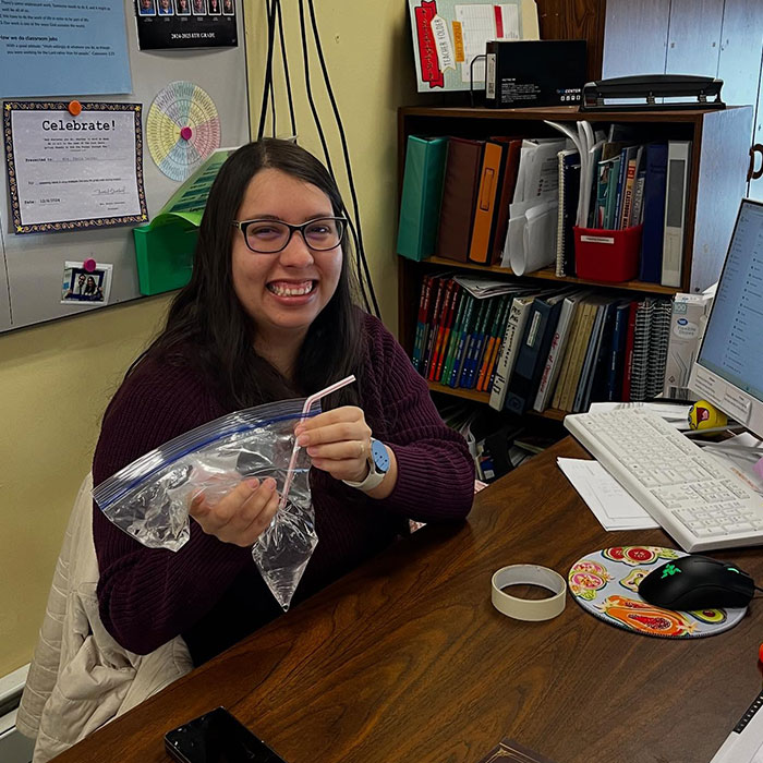 Woman smiling at desk drinking from a plastic bag with a straw, showcasing creative sippers using anything but a cup.