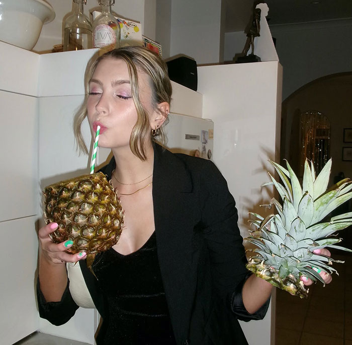 Woman sipping from a hollowed-out pineapple with a straw, showcasing creative sippers using anything but a cup.