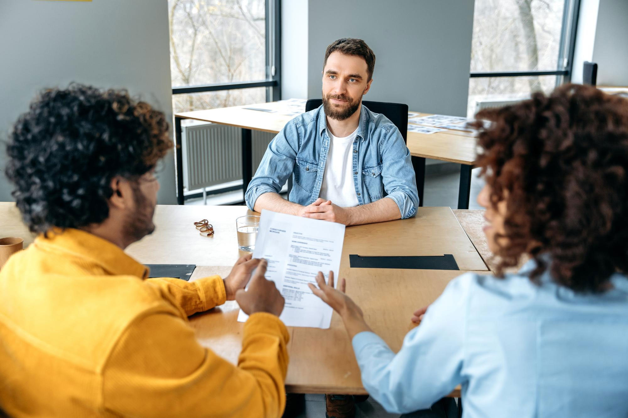 Man in denim shirt sitting in an interview setting with two people reviewing a resume discussing career and job decisions. - 6