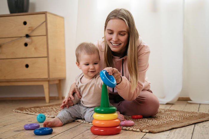 Young nanny playing with baby using colorful stacking rings while sitting on a rug in a cozy room