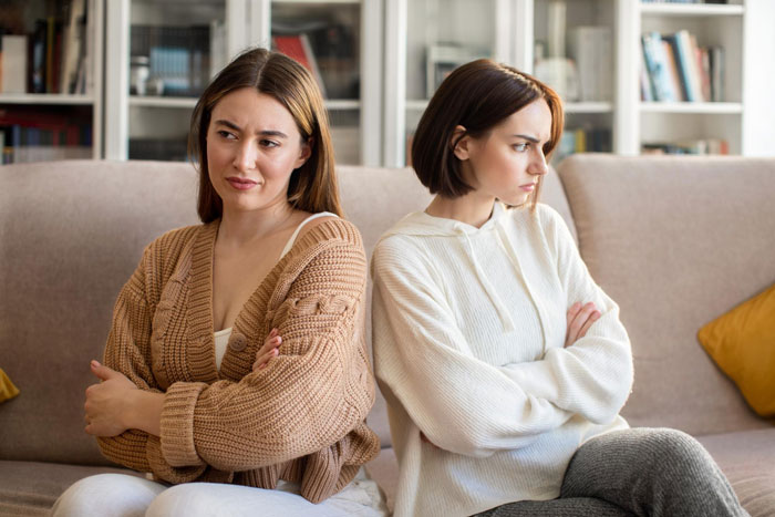 Two women sitting on a couch with crossed arms, showing tension related to nanny working long hours and family demands.