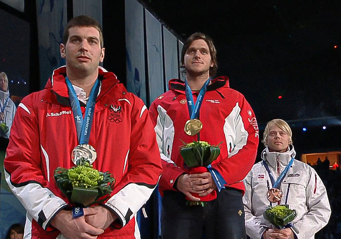 Three Olympic medalist skiers wearing medals and holding flowers during a podium ceremony at a nighttime event. - 14