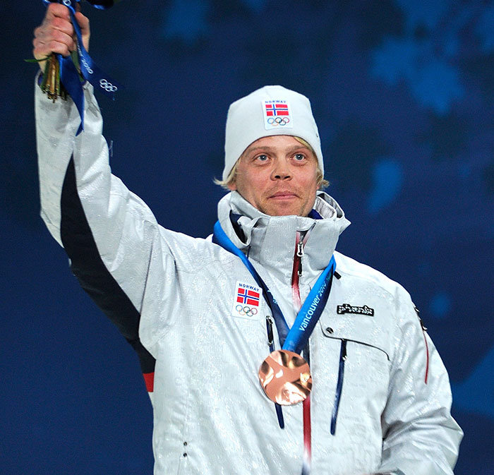 Norwegian Olympic medalist skier wearing white jacket and hat, holding up medal in a celebratory pose. - 1