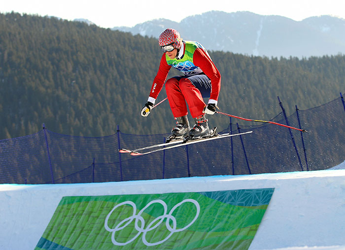 Olympic medalist skier in mid-air during a jump at a snowy mountain slope with Olympic rings banner below. - 3