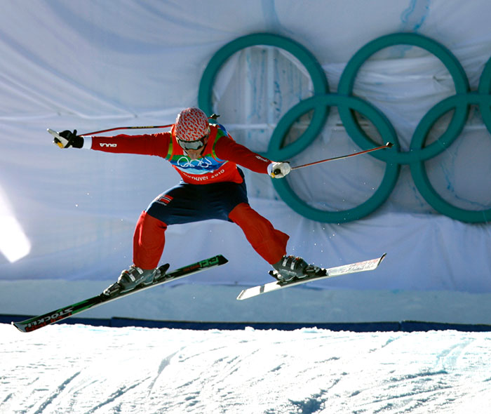 Olympic medalist skier wearing red outfit performing a jump with Olympic rings in background on a snowy slope. - 6
