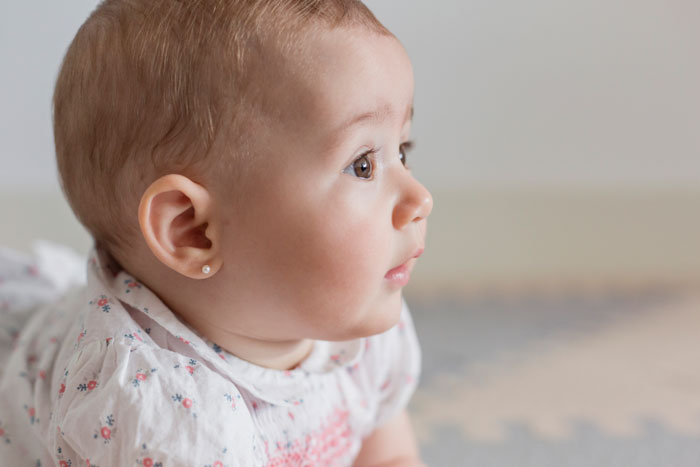 Baby with gold stud earrings after unlicensed ear piercing during a family visit, mom visibly upset in a home setting.