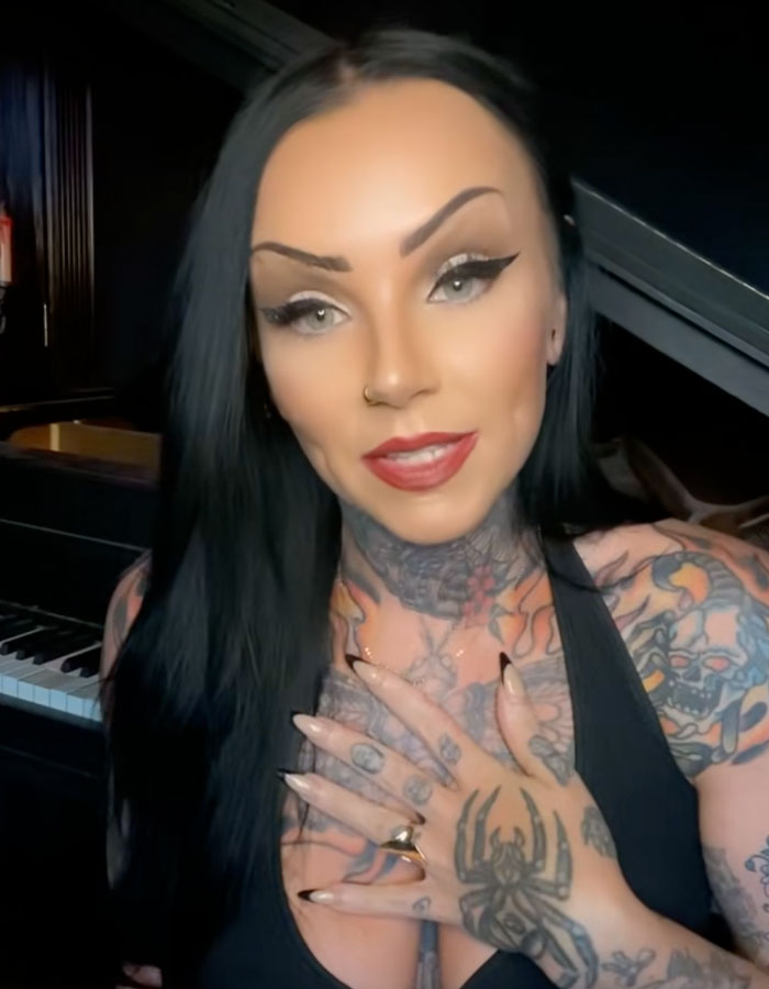 Young woman with tattoos, wearing a black top, sharing insights about autopsy tech and strange ways people have passed away.