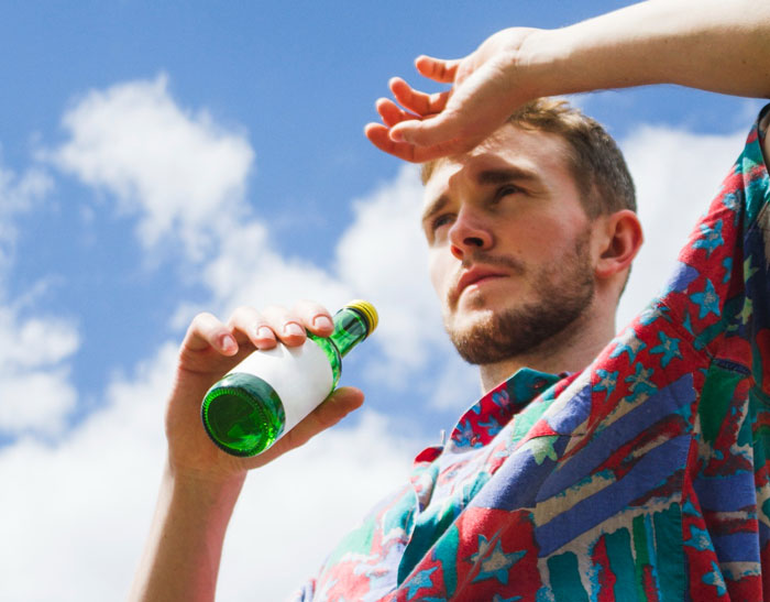 Man shielding eyes from sun while holding a green bottle, highlighting warnings about alcohol during extreme heat in 18 states. - 1