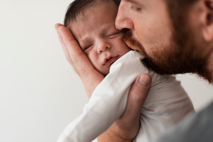 Father carrying infant by the arms while gently wiping the baby's face with a dishrag, unaware of wife's concern.