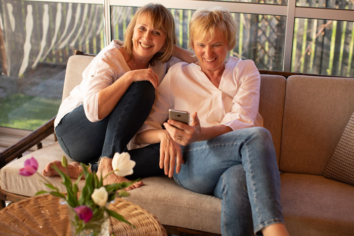 Two women laughing and looking at a phone, enjoying a moment in a cozy sunlit room. Two women laughing and looking at a phone, enjoying a moment in a cozy sunlit room.