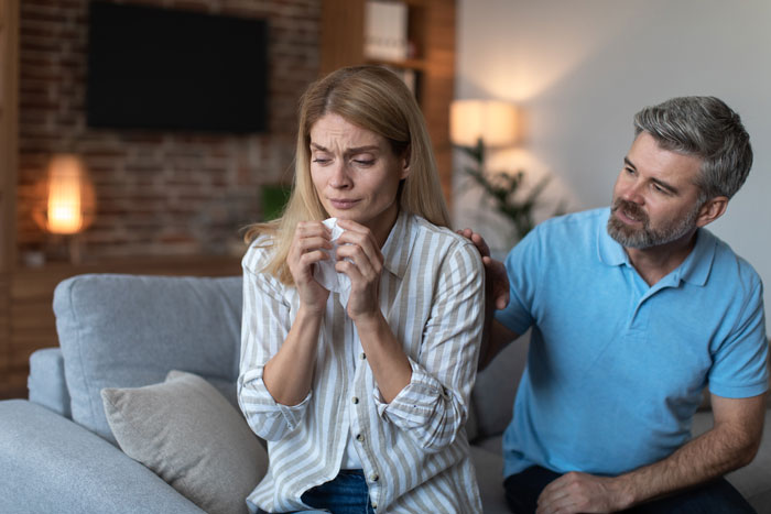 Woman upset on couch holding tissue while man comforts her during a tense family discussion about babysitting step-siblings.