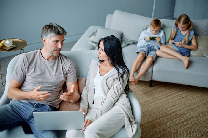 Parents having a serious discussion on couch while two children ignore them, illustrating babysitting step-siblings challenge.