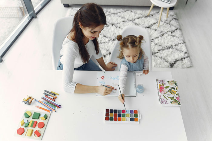 Woman babysitting young girl with paints and crayons at white table in bright living room
