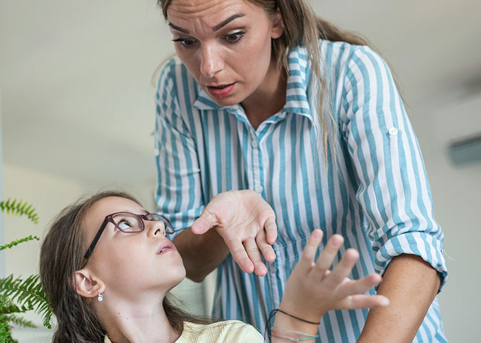 Woman in striped shirt angrily gesturing while teen girl with glasses looks up during a tense family moment. Woman in striped shirt angrily gesturing while teen girl with glasses looks up during a tense family moment.