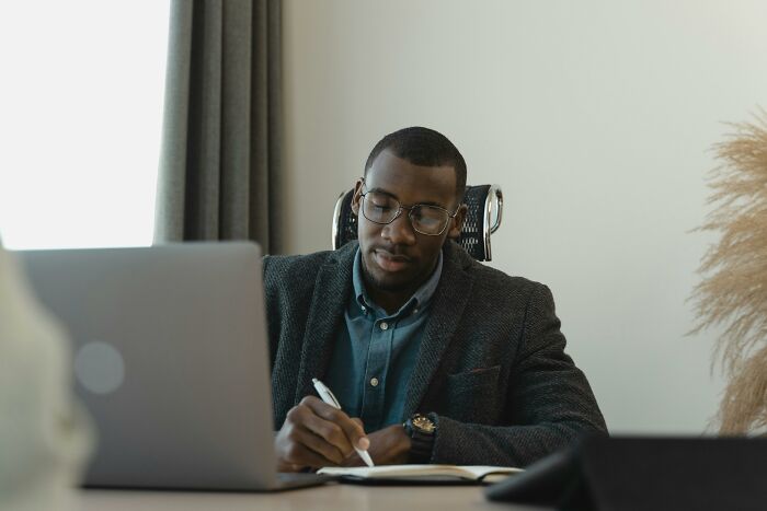 Man wearing glasses and a blazer working on a laptop and writing notes, showing bare minimum or princess treatment concept.
