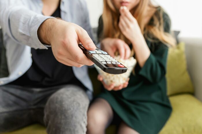 Couple on a couch sharing popcorn while one person holds a remote control deciding bare minimum or princess treatment.