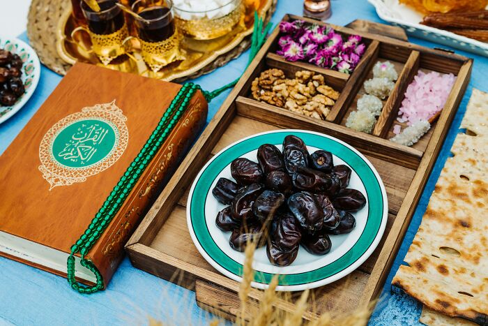 Wooden tray with dates, nuts, and spices next to a Quran book and prayer beads on a blue cloth background.