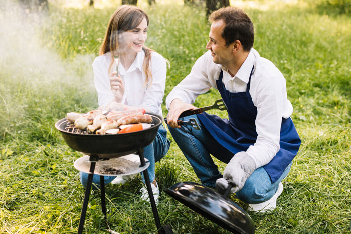 Couple enjoying outdoor barbecue, with man grilling and woman holding fork, hinting at a shocked bestie and possible hidden affair.