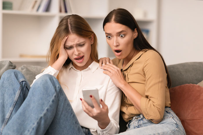 Two young women, one upset and the other shocked, looking at a smartphone, reflecting influencer drama and studio party conflict.
