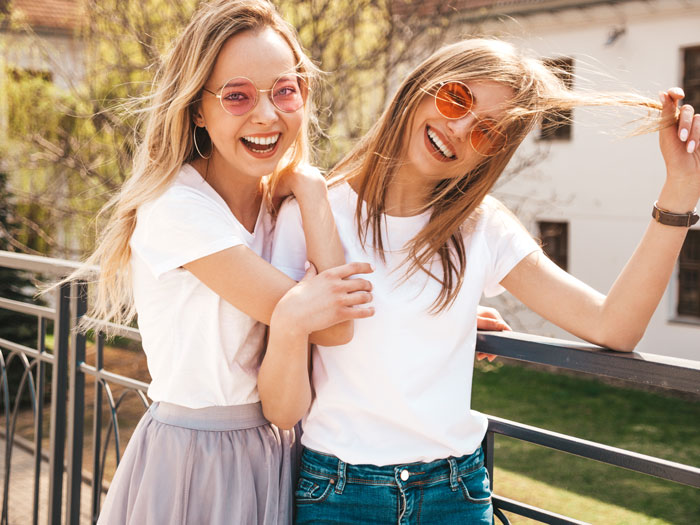 Two young women wearing sunglasses and white shirts, smiling and posing together outdoors on a balcony.