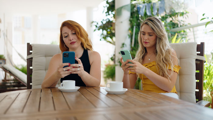 Two women sitting apart at a table, both focused on their phones, reflecting tension between bestie and wannabe influencer.