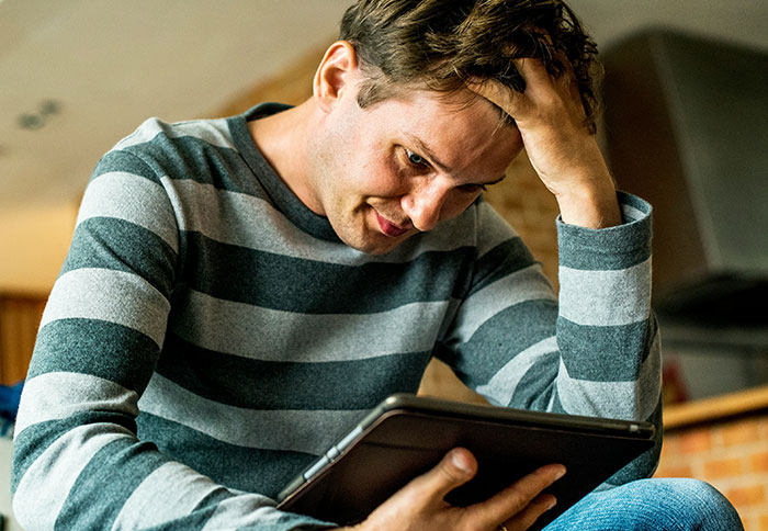 Young man in a striped shirt looking stressed while checking email on a tablet, best friend saw girlfriend with another man.