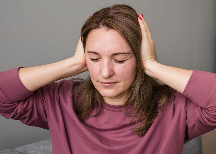 Woman in a maroon sweater covering her ears with hands, expressing stress while sitting against a gray background. - 40