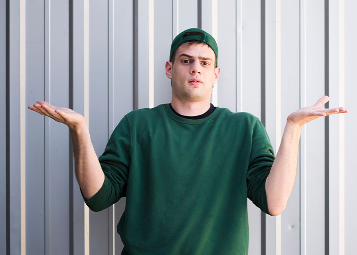 Young man in green sweatshirt and cap shrugging with hands up against a gray wall, illustrating life cheat codes concept. - 8