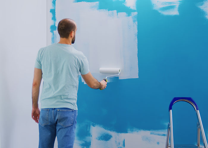 Man using a paint roller to apply light blue paint over dark blue wall showing life cheat codes in home improvement. - 21
