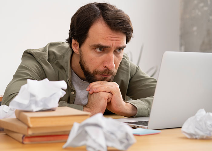 Man focused on laptop screen surrounded by crumpled papers and books, illustrating life cheat codes concept. - 31