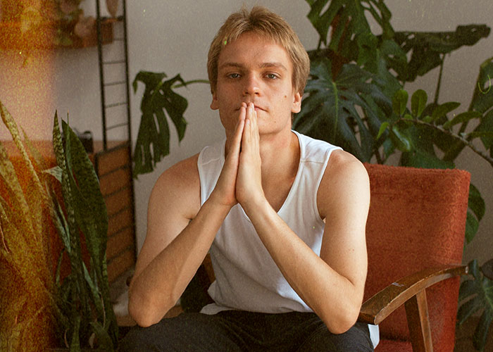 Young man in a white tank top sitting thoughtfully in a living room surrounded by plants, representing life cheat codes. - 13