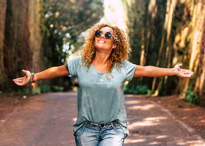 Happy woman wearing sunglasses with arms outstretched on tree-lined path, enjoying life and its cheat codes outdoors. - 4