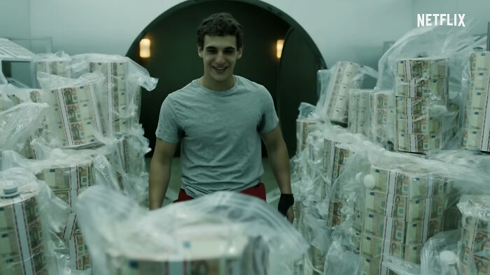 Young man smiling amid stacks of wrapped money bundles in a scene from one of the best Netflix shows. - 14