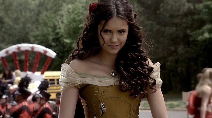 Young woman with long curly hair in a vintage dress at an outdoor event with people and a band in the background. - 23