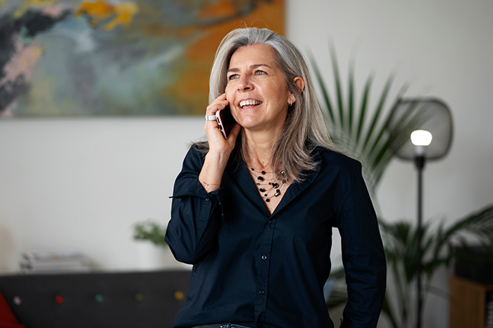 Mature woman with gray hair smiling while talking on phone in a modern living room with plants and artwork.