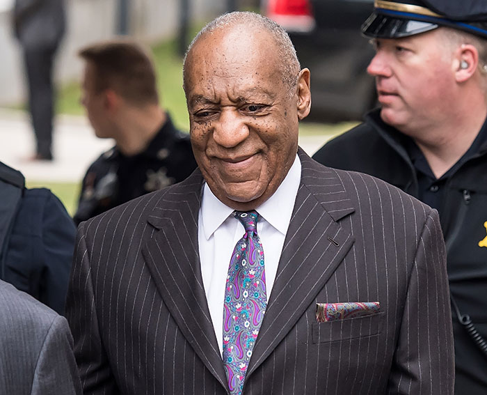 Bill Cosby wearing a pinstripe suit and patterned tie, surrounded by police officers outdoors. - 1