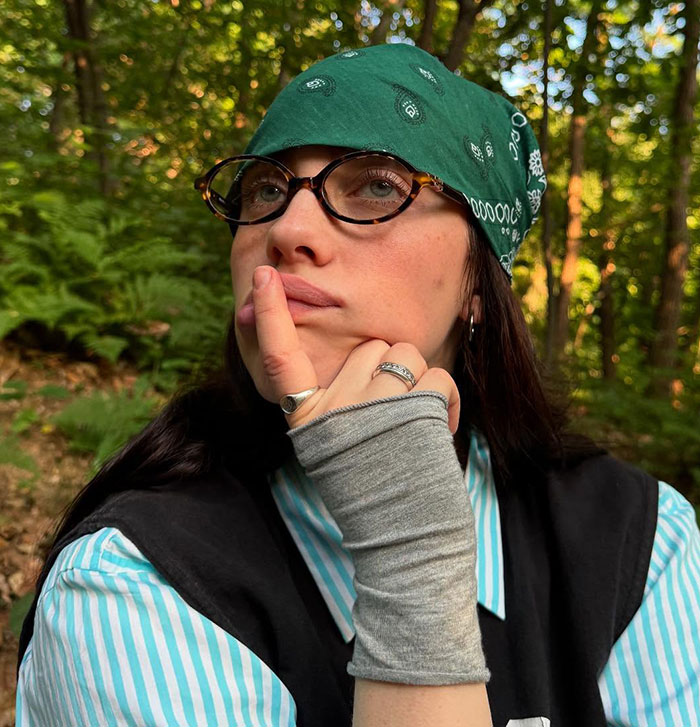 Billie Eilish wearing glasses and green bandana, posing thoughtfully outdoors with trees in the background