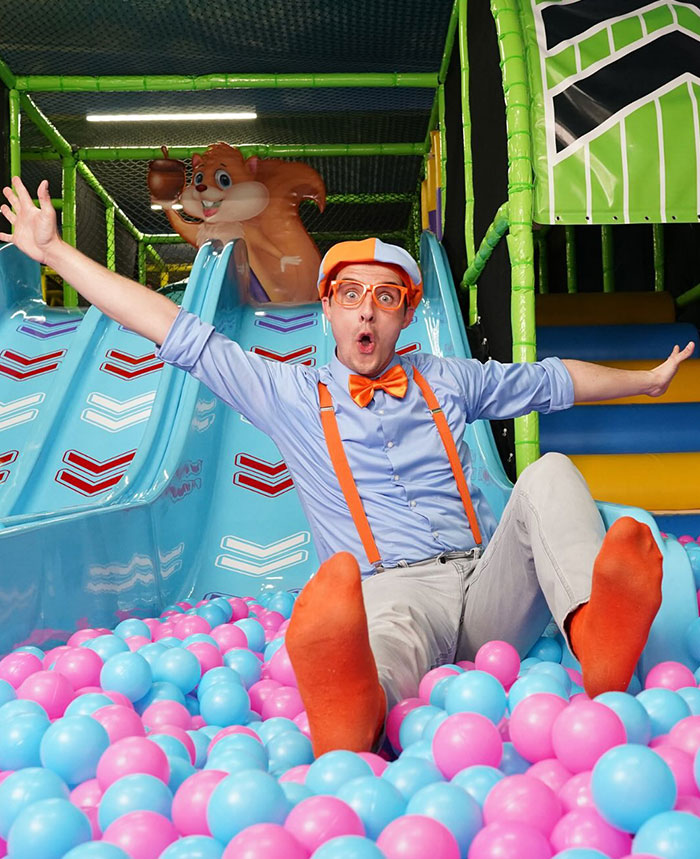 Man dressed as Blippi with orange suspenders and glasses playing in a colorful kids’ ball pit and slide area. - 5