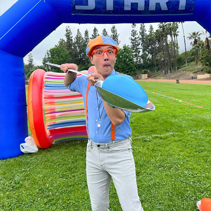 Blippi wearing orange glasses and hat, holding a shiny spoon and blue bowl at an outdoor kids’ event start line. - 11