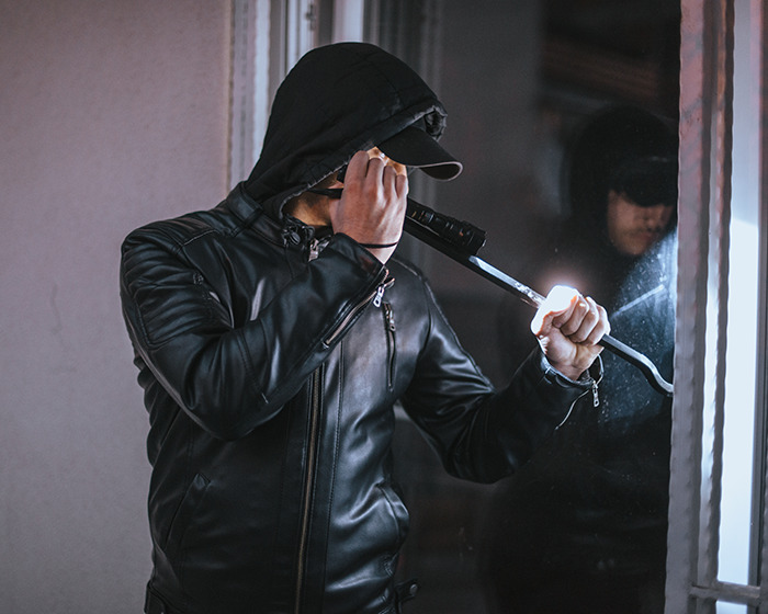 Person wearing a black leather jacket and hood using a crowbar to break into a house, illustrating risks of blurring houses on Google Maps.